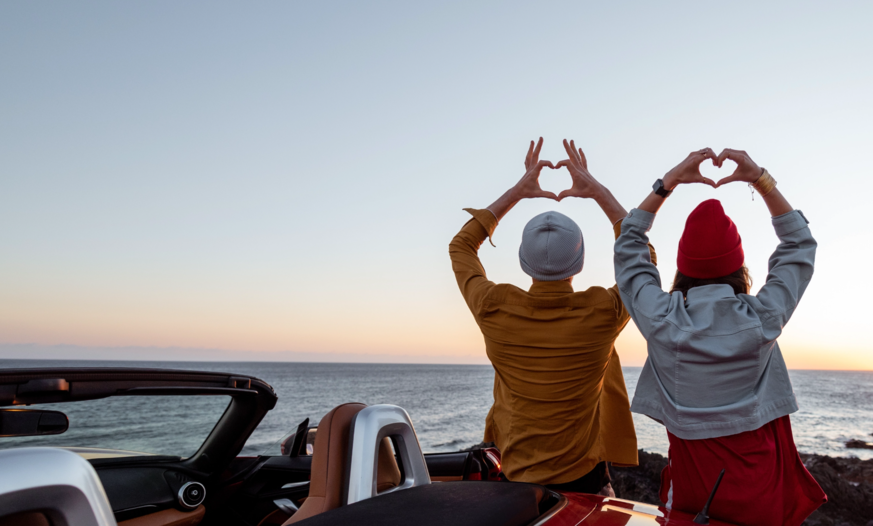 Mädchen und Junge von hinten formen Herz mit Händen Mädchen und Junge von hinten sitzen auf Auto bei Sonnenuntergang und formen Herz mit Händen