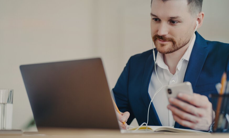 Man vor Laptop mit Handy in der Hand und Kopfhörer im Ohr