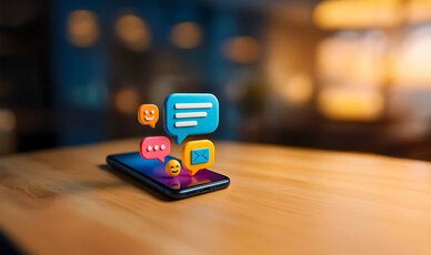 Smartphone liegt auf dem Tisch. Bunte 3D-Emojis schweben heraus. 